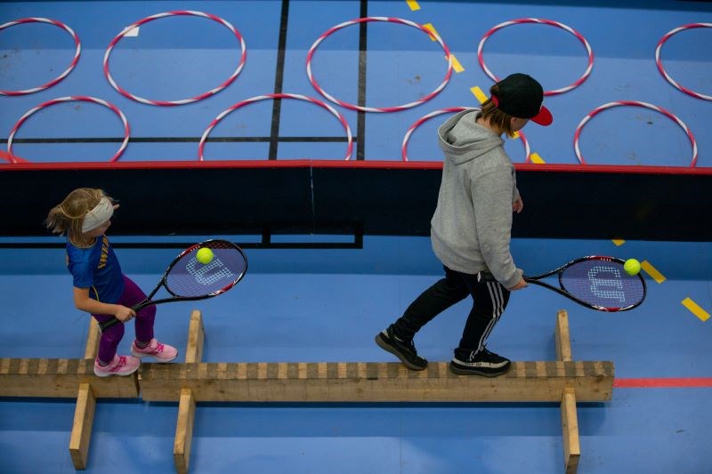 Två personer med tennisracket balanserar på ett redskap i en idrottshall